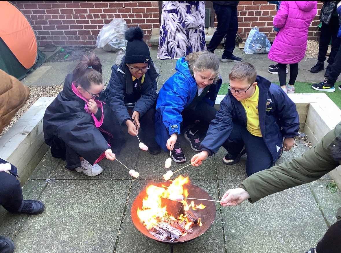 Children gathered around a small outdoor fire pit roasting marshmallows on sticks in a paved courtyard.