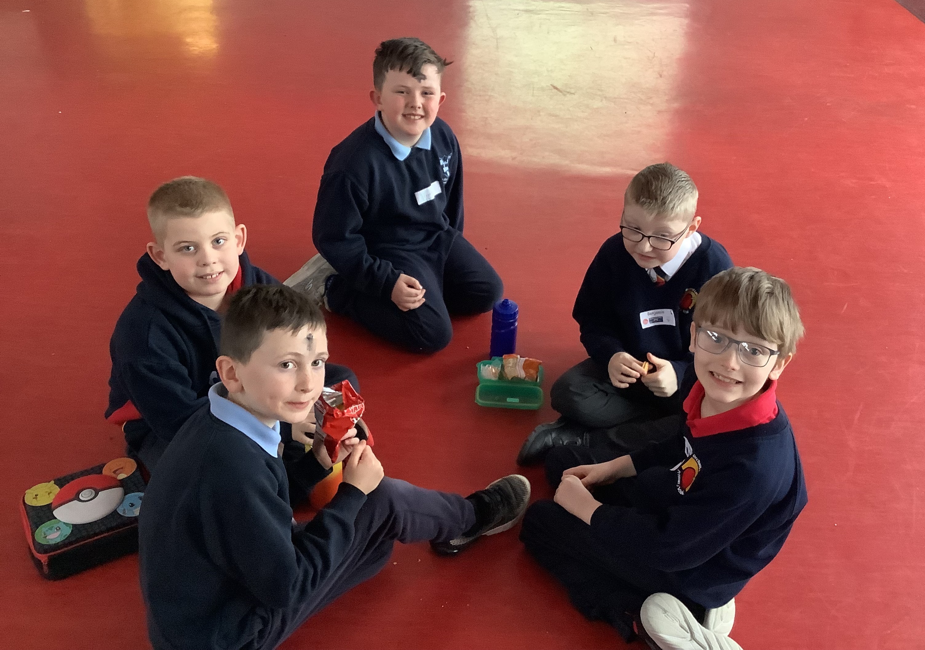Five young boys sitting in a circle on a red floor, sharing snacks and drinks during a break at school.