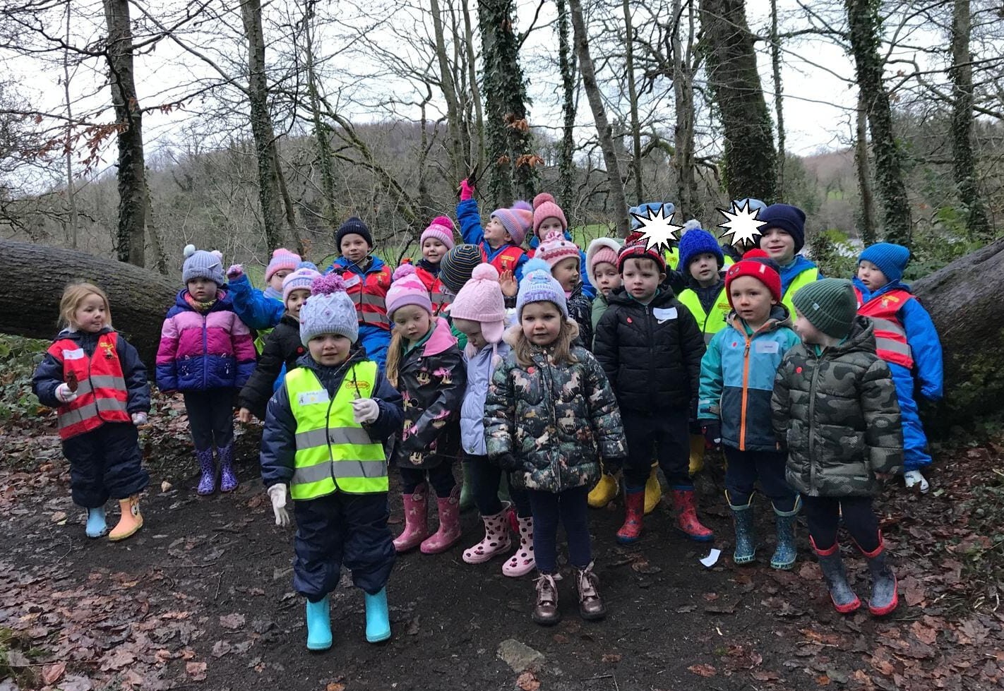 Group of young children dressed in colourful winter jackets and hats standing together in a leaf-covered forest clearing.