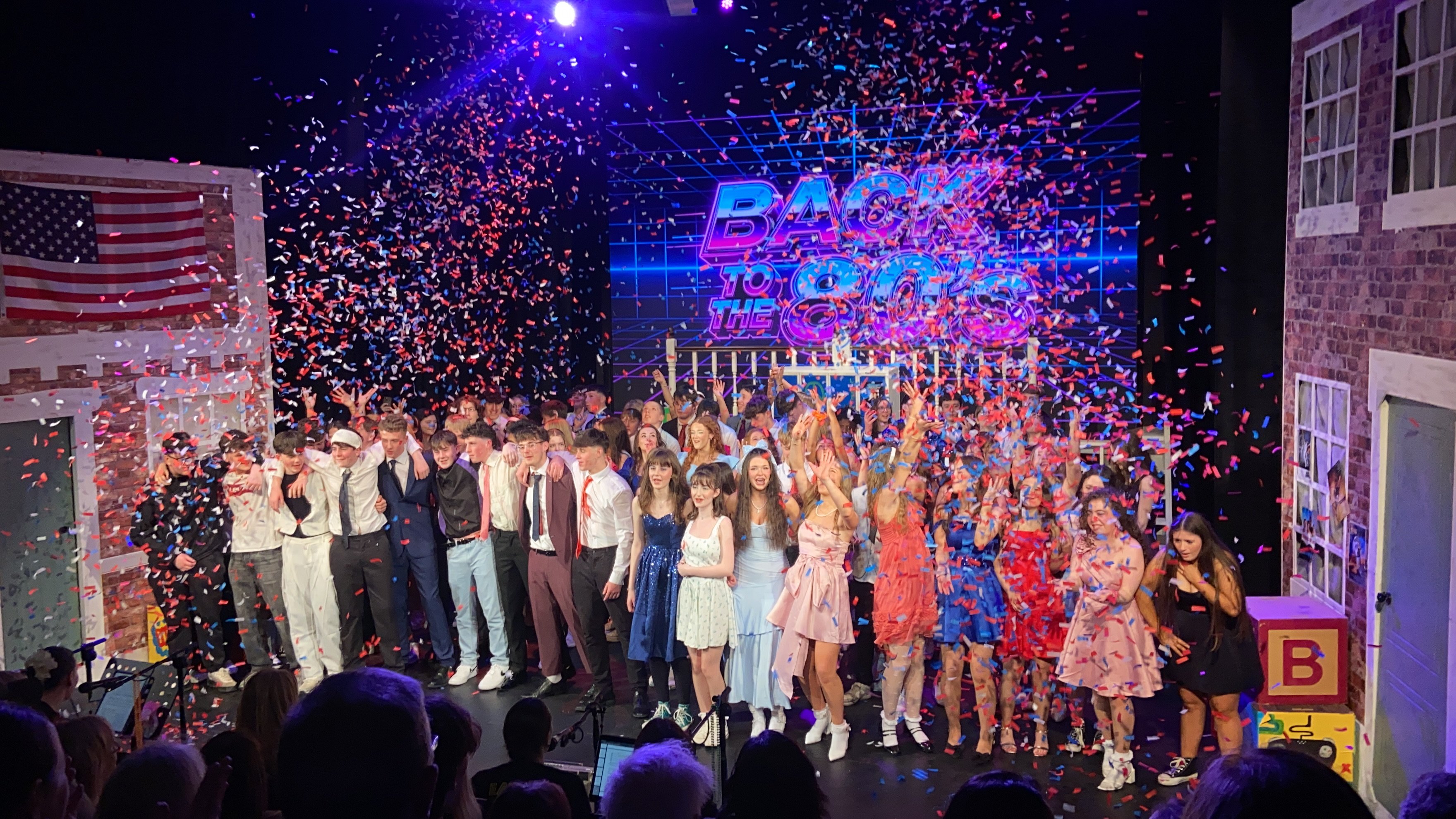 Large group of young performers on stage celebrating with confetti falling during a Back to the 80s musical show.   